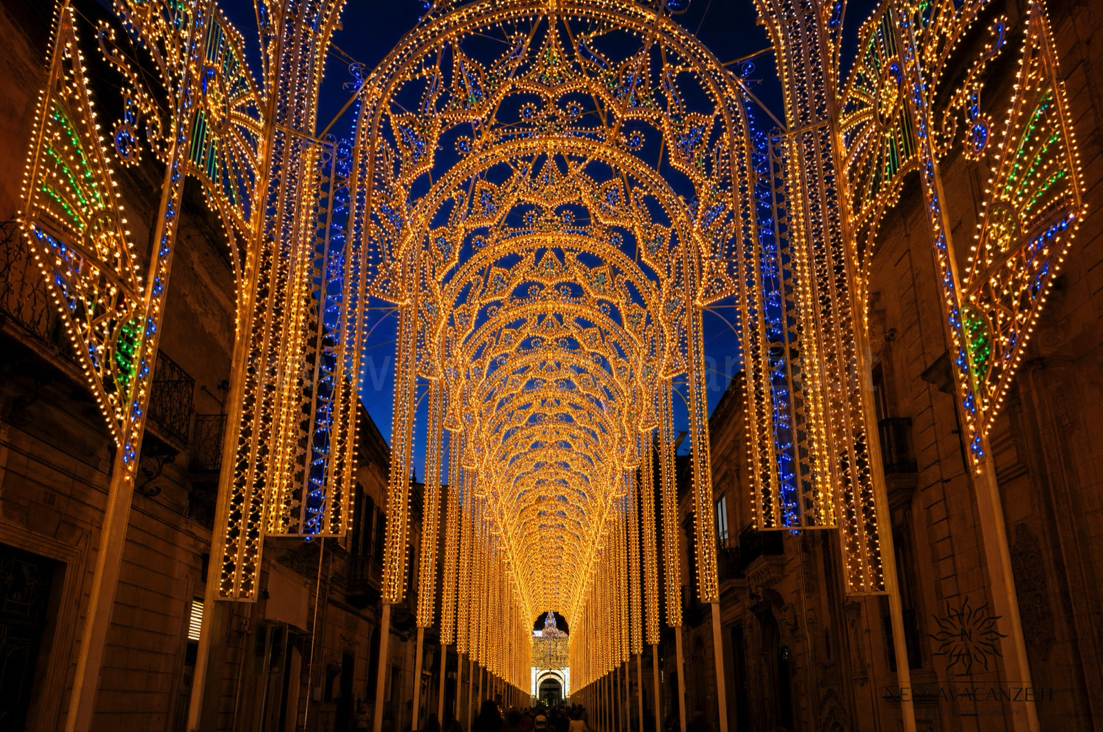 Luminarie salentine sopra la via di un paese in festa: archi di legno illuminati da migliaia di lampadine, come per San Nicola a Salve