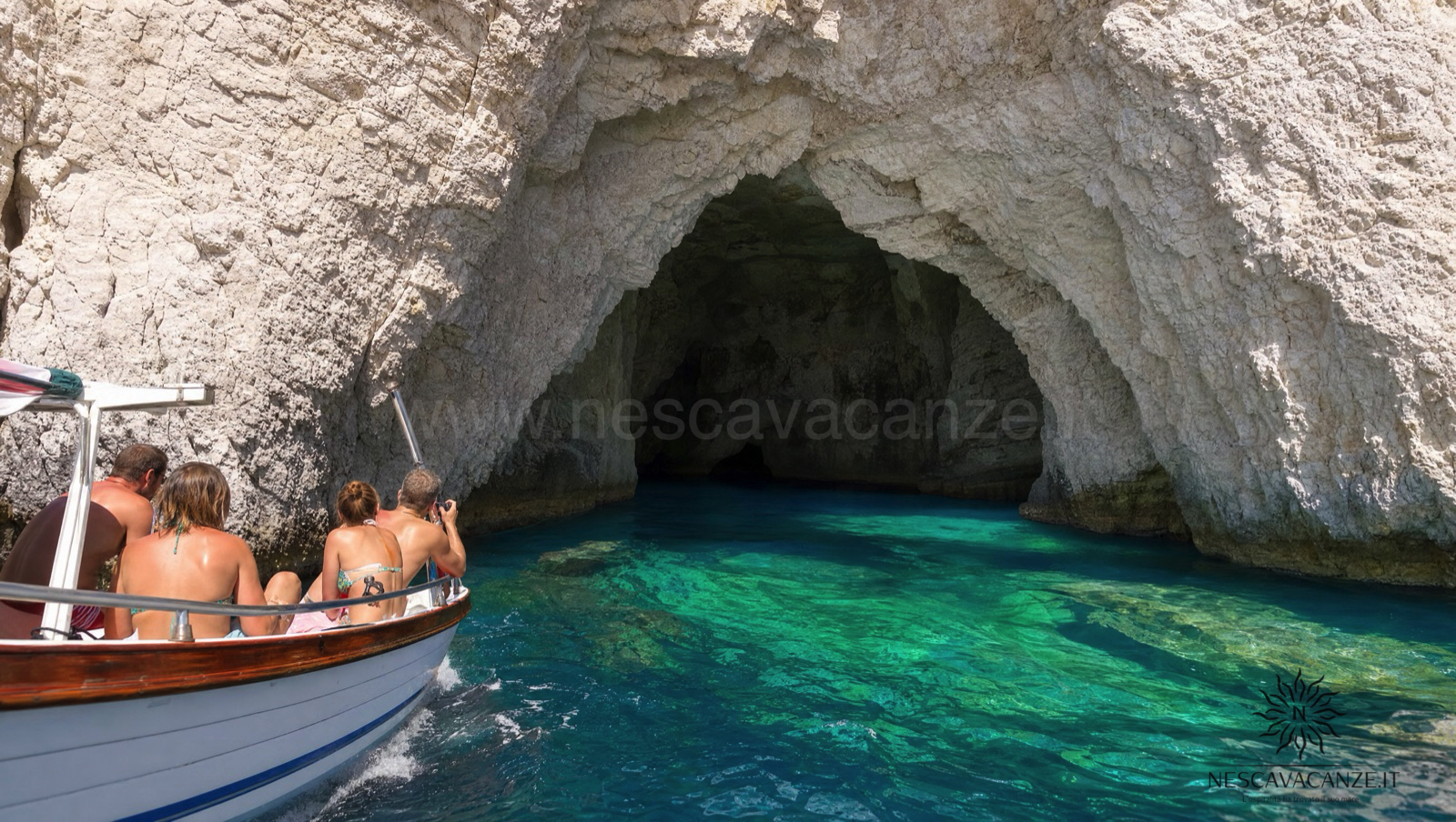 Piccola barca con passeggeri all'ingresso di una grotta marina dalla volta di roccia bianca, con l'acqua turchese che riflette la luce, lungo la costa del Salento