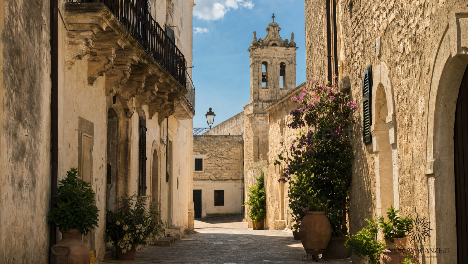 Vicolo di pietra di un borgo dell'entroterra salentino come Specchia, con case in pietra chiara, un campanile barocco, bouganville e piante in vaso