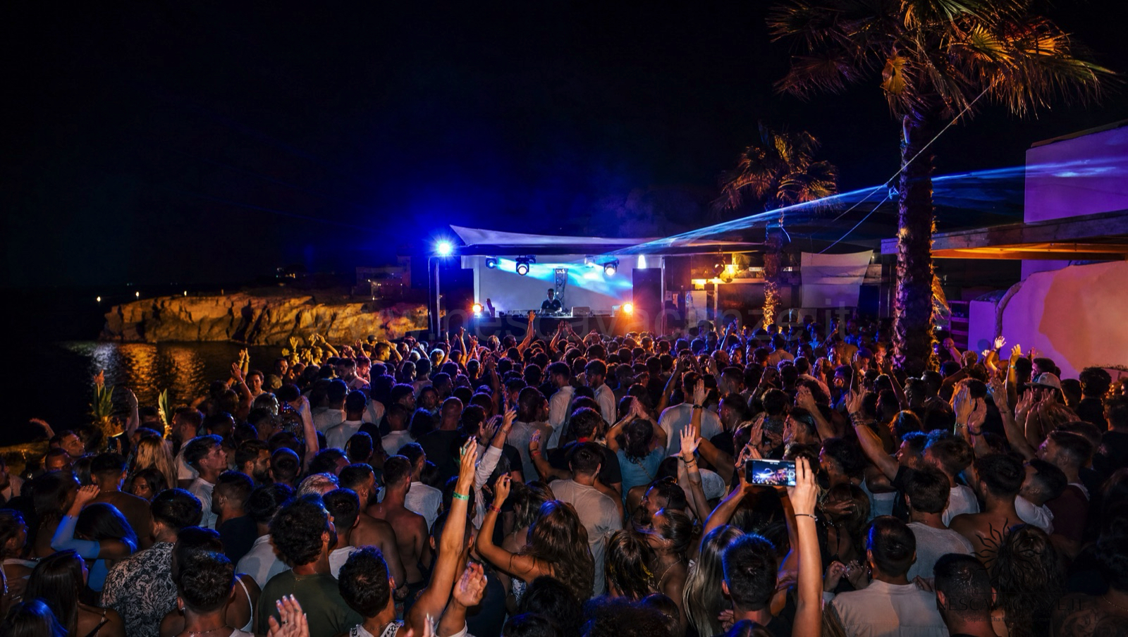 Folla che balla in un beach club all'aperto sul mare di notte con luci blu e palme nel Salento